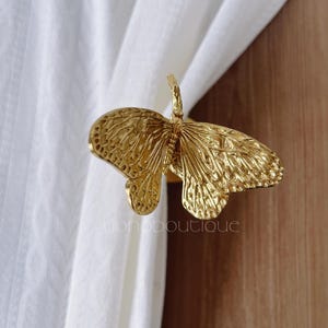 May include: A gold-colored butterfly curtain tieback. The butterfly has detailed wing patterns and is attached to a curved stem. The tieback is holding a white curtain. The text "yonoboutique" is visible on the bottom right.