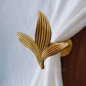 May include: A gold-coloured, leaf-shaped curtain tieback. The metal tieback features a detailed, ribbed design. It is holding a white curtain. The text "yoneboutique" is visible on the bottom right of the image.