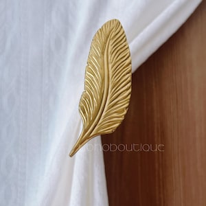 Könnte beinhalten: Ein goldfarbener, federförmiger Vorhanghalter. Der Vorhanghalter ist aus Metall gefertigt und hat detaillierte Adern. Die Feder ist an einem weißen Vorhang befestigt. Das Wort "ohoboutique" ist auf dem Vorhang aufgedruckt.