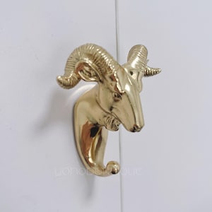 以下が含まれることがあります： 白い表面に取り付けられた、金色の羊の頭の形をしたフック。 フックは、詳細な角と下の湾曲したフックを備えた羊の頭の形をしています。 フックはおそらく金属製で、装飾品です。