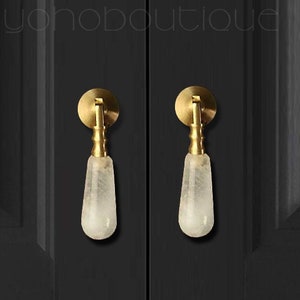 Pode incluir: Duas puxadores de armário com base circular dourada e uma pega em forma de gota de vidro fosco transparente. Os puxadores são montados numa porta de armário preta. O texto "yohoboutique" é visível no fundo.