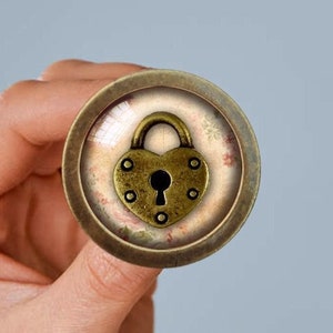 May include: A round, gold-toned metal button with a heart-shaped padlock design. The padlock is gold with a keyhole and is set against a background of pink and white floral patterns.