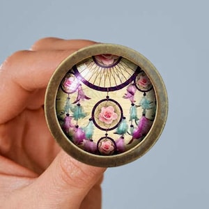 Op de afbeelding: Een ronde, bronzen hanger met een glazen inzetstuk met een kleurrijk droomvangerontwerp met roze bloemen en paarse veren.