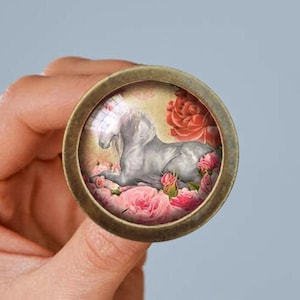 May include: A round, antique brass cabinet knob featuring a white unicorn resting among pink roses. The image is set within a clear glass dome. The knob has a vintage, romantic aesthetic, suitable for furniture or decor.