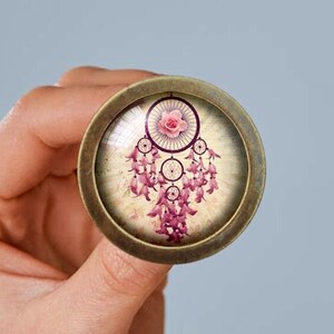 Pode incluir: Um pingente redondo de cor bronze com um inserto de vidro que apresenta um apanhador de sonhos rosa e branco com uma rosa rosa no topo.