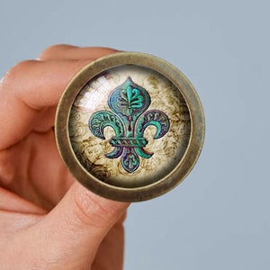 Może przedstawiać: Okrągły, brązowy guzik metalowy z szklanym wierzchem, przedstawiający zielony i fioletowy wzór lilii na kremowym tle.
