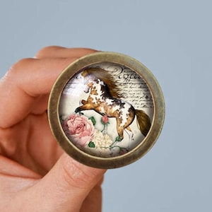 May include: A round, antique gold-toned metal button with a glass cabochon featuring a watercolour painting of a brown and white spotted horse running with a pink rose in the foreground. The background is a vintage-style cream paper with cursive writing.