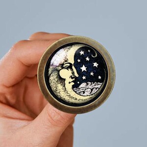 May include: A round, brass-colored pin with a black and white image of a crescent moon with a face and stars. The moon is on a blue background.
