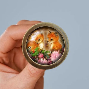Könnte beinhalten: Eine runde, bronzefarbene Anstecknadel mit einer Glaskuppel, die ein Gemälde von zwei roten Füchsen mit rosa Blumen zeigt.