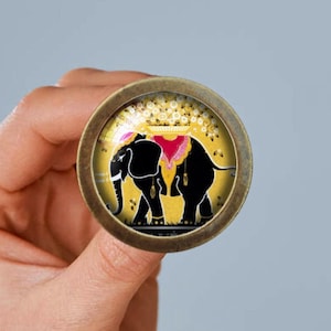 Pode incluir: Um puxador de armário redondo, cor bronze, com um design decorativo de elefante. O elefante é preto com detalhes dourados e uma sela rosa, sobre um fundo amarelo. O puxador é segurado em uma mão.