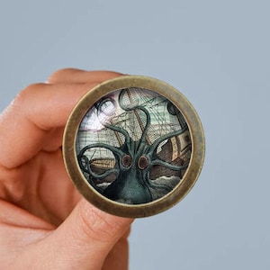 May include: A round, brass-colored pin with a glass dome featuring a vintage illustration of a giant octopus attacking a ship. The octopus is green and has large, menacing eyes.
