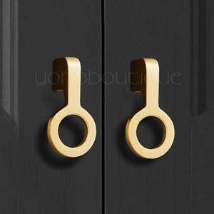 May include: Two gold-colored cabinet pulls with a unique circular design. The pulls are mounted on a dark gray cabinet door. The pulls have a modern, minimalist aesthetic.