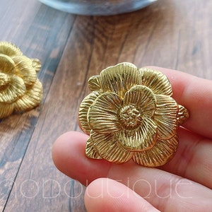 Può includere: Un paio di orecchini dorati a forma di fiore. Gli orecchini sono in metallo e hanno una superficie testurizzata.