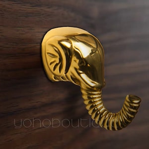 Op de afbeelding: Een gouden, olifantvormige wandhaak met een gekrulde slurf. De haak is gemonteerd op een donker houten oppervlak.