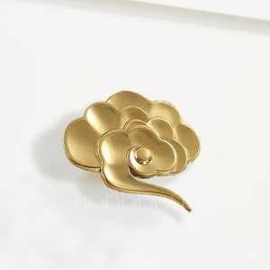 Può includere: Pomo per mobile in metallo dorato a forma di nuvola stilizzata. Il pomo ha una superficie liscia e arrotondata e una coda leggermente curva.