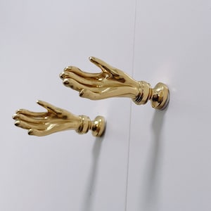 May include: Two gold-toned metal hand-shaped wall hooks. The hands are outstretched with palms facing upwards.