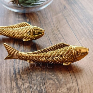 May include: Two gold-colored fish figurines on a wooden surface. The fish are detailed with scales and have a realistic appearance.