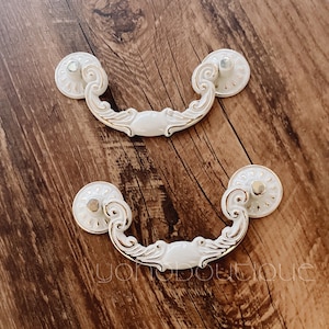 May include: Two white ornate drawer pulls with gold accents. The pulls are shaped like a traditional handle with a rounded top and a decorative scroll design.