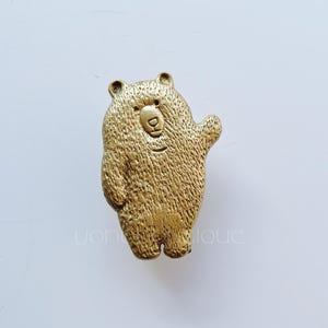 Pode incluir: Uma figura de urso dourada com uma superfície texturizada. O urso está em pé sobre suas patas traseiras e tem sua pata direita levantada em um aceno.