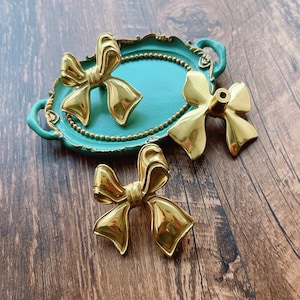 May include: Three gold-coloured bow-shaped drawer pulls on a teal green tray with a beaded edge.