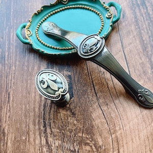 Könnte beinhalten: Zwei antike Schrankknöpfe mit einem Fleur-de-lis-Design. Ein Knopf ist silberfarben und der andere ist messingfarben.