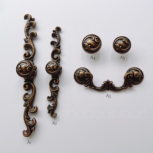 May include: Five antique brass cabinet knobs and pulls with ornate designs. The knobs are round with a raised centre and the pulls are shaped like a curved handle with a decorative end.