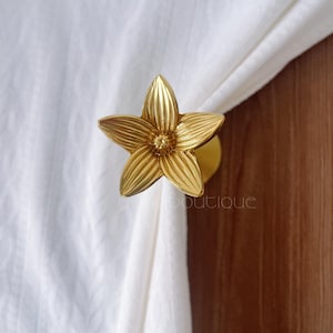 Peut inclure: Un embrasse-rideau doré en forme de fleur. L'embrasse-rideau est en métal et présente un motif floral détaillé. L'embrasse-rideau maintient un rideau blanc. Le mot "boutique" est visible dans l'image.