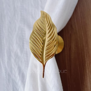 May include: A gold-coloured leaf-shaped curtain tieback. The tieback is made of metal and has detailed veining. The leaf is attached to a round base. The tieback is holding a white curtain. The text "yono boutique" is visible.
