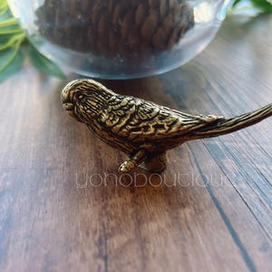 May include: A detailed, golden-colored bird figurine. The bird is perched on a small base, with textured feathers and a long tail. The figurine is likely made of metal and is a decorative item.