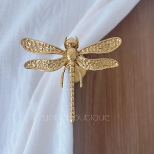 Puede incluir: Un adorno decorativo dorado con forma de libélula. La libélula tiene alas y cuerpo detallados, y está montada sobre una base circular. La imagen está colocada sobre una tela blanca y una superficie de madera marrón.