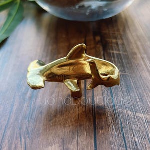 Könnte beinhalten: Ein goldfarbener Ring in Form eines Hammerhais. Der Ring besteht aus Metall und hat ein detailliertes Design, einschließlich Kopf, Körper und Flossen des Hais. Der Ring wird auf einer Holzoberfläche präsentiert.