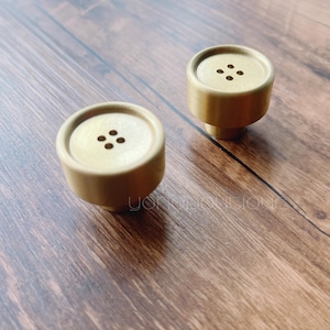 May include: Two gold-toned metal cabinet knobs with a round shape and four holes in the center. The knobs are on a wooden surface.