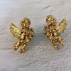 以下が含まれることがあります： 詳細な翼と巻き毛を持つ、2つの金色のケルビムの置物。ケルビムは座った姿勢で、腕を上げています。背景は、テクスチャ加工されたオフホワイトの表面です。これらの装飾的な置物は、おそらく室内装飾に使用されます。