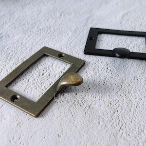 May include: Two rectangular label holders, one in antique brass and the other in black. The brass holder has a curved handle, while the black one has a small, round knob. Both have screw holes for mounting.