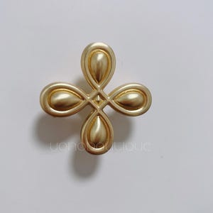May include: A gold-colored, four-petal cabinet knob with a matte finish. The knob has a cross-like design with rounded petal shapes and a square center. The image is of the knob against a white background.