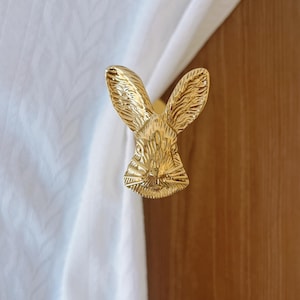 May include: A gold-coloured bunny napkin ring. The bunny's head is detailed with textured fur and long ears. The napkin ring is holding a white cloth. The background is a wooden surface.