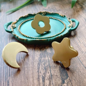 以下が含まれることがあります： 金色の装飾的な引き出しノブのコレクション。花の形、三日月、星の形です。花のノブは、金の縁取りのあるターコイズ色のトレイに置かれています。他のノブは木製の表面にあります。