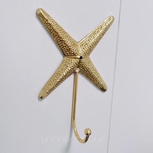 Könnte beinhalten: Ein goldfarbener Wandhaken in Form eines Seesterns. Der Haken besteht aus Metall und hat eine strukturierte Oberfläche. Der Haken ist an einem langen, gebogenen Stiel mit einer kleinen Kugel am Ende befestigt. Der Haken ist auf einer weißen Oberfläche montiert.