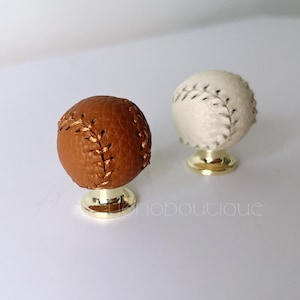 May include: Two baseball-themed cabinet knobs. One is brown with gold stitching, the other is white with black stitching. Both knobs have a gold-coloured base. The knobs are likely for a sports-themed room.