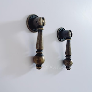 Puede incluir: Dos tiradores de armario de latón antiguo con un diseño redondeado y ornamentado. Los tiradores están unidos a una superficie blanca.