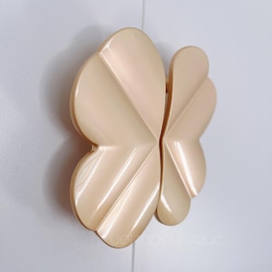 Pode incluir: Um par de puxadores decorativos para armários com forma de borboleta. Os puxadores são feitos de plástico brilhante e perolado e têm uma cor bege claro.