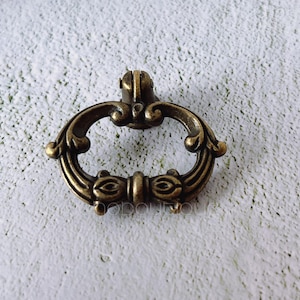 Pode incluir: Um puxador de gaveta de latão em estilo antigo com um design oval ornamentado. O puxador apresenta redemoinhos decorativos e um detalhe central em forma de olho. A ferragem tem uma cor de latão envelhecido e quente.