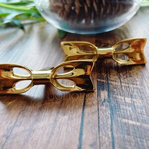 May include: Two gold-coloured bow-shaped hair clips. The clips have a polished, reflective surface and a cutout design in the centre. They are placed on a wooden surface, with a blurred background that includes greenery and a glass container.