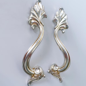 Può includere: Due pomelli per cassetti argentati e ornati, con un design a forma di foglia nella parte superiore e una maniglia curva a forma di voluta. I pomelli hanno un'estetica vintage, con sottili accenti dorati e una superficie strutturata dettagliata.