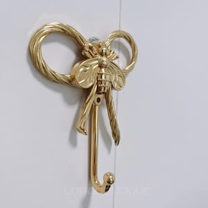 以下が含まれることがあります： 金色の壁掛けフック、蜂のデザイン。フックは装飾的なリボン型のトップと、中央に様式化された蜂が特徴です。フックは下方に伸び、アイテムを吊るすための湾曲した端があります。フックは白い表面に取り付けられています。