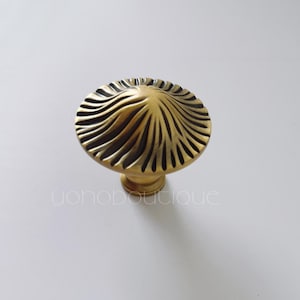 Pode incluir: Um puxador de armário dourado com um design detalhado e ondulado na cabeça redonda. O puxador tem uma base curta e cilíndrica. O texto "yohoboutique" é visível na parte inferior da imagem.