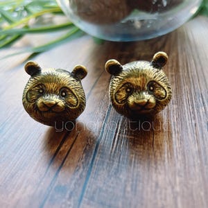 Pode incluir: Dois brincos de pressão com cabeça de panda em tom bronze. Os brincos apresentam detalhes, incluindo orelhas pequenas e expressões faciais. Estão sobre uma superfície de madeira.