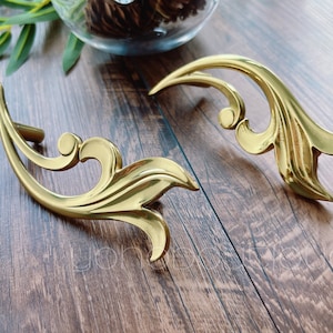 Puede incluir: Dos tiradores de cajón de metal dorado adornados con un diseño de hoja decorativo.