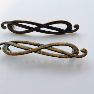 Op de afbeelding: Twee antieke, goudkleurige metalen ladegrepen met een ingewikkeld, verweven ontwerp. De grepen zijn in de vorm van een acht.