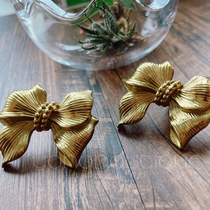 Puede incluir: Un par de pendientes de color dorado en forma de lazo. Los pendientes están hechos de metal y tienen un acabado texturizado.